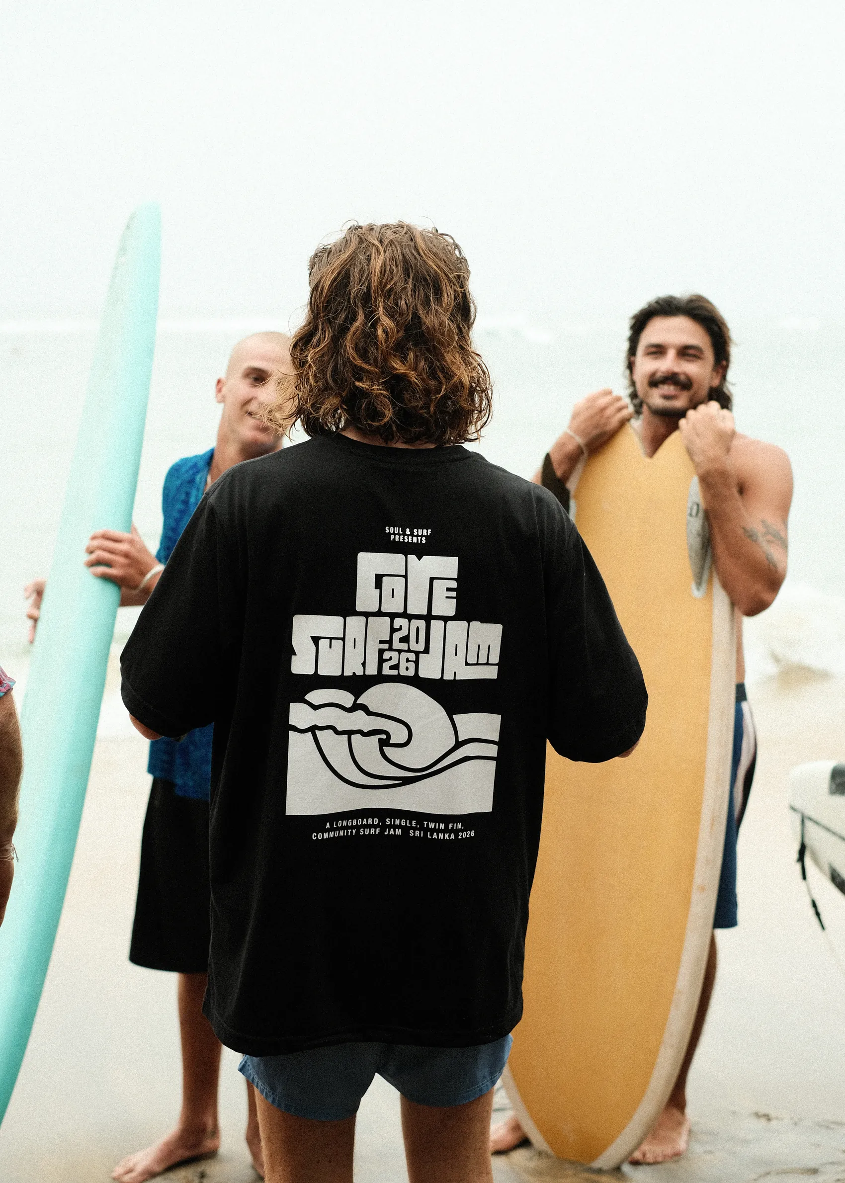 Surfer on the beach in the Cove Surf Jam 2026 T-shirt, friends holding boards either side