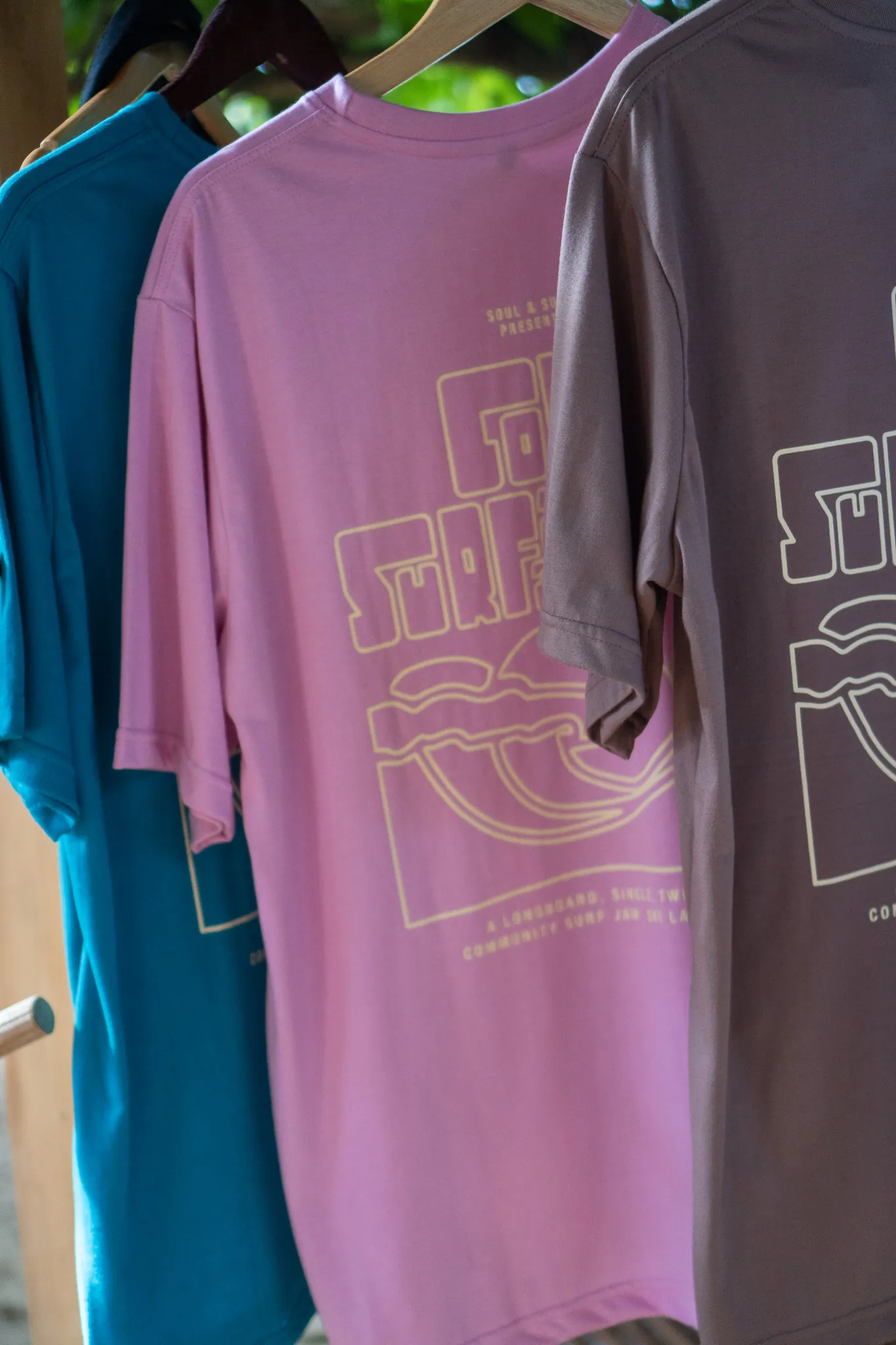 Cove Surf Jam T-shirts in blue, pink and grey hanging on wooden hangers